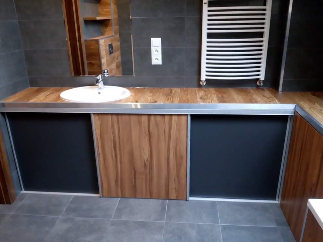 Nowoczesna łazienka z szarymi płytkami, drewnianym blatem, umywalką nablatową i grzejnikiem drabinkowym; szafki pod umywalką z przesuwnymi drzwiami z elementami drewnopodobnymi i czarnymi wstawkami.