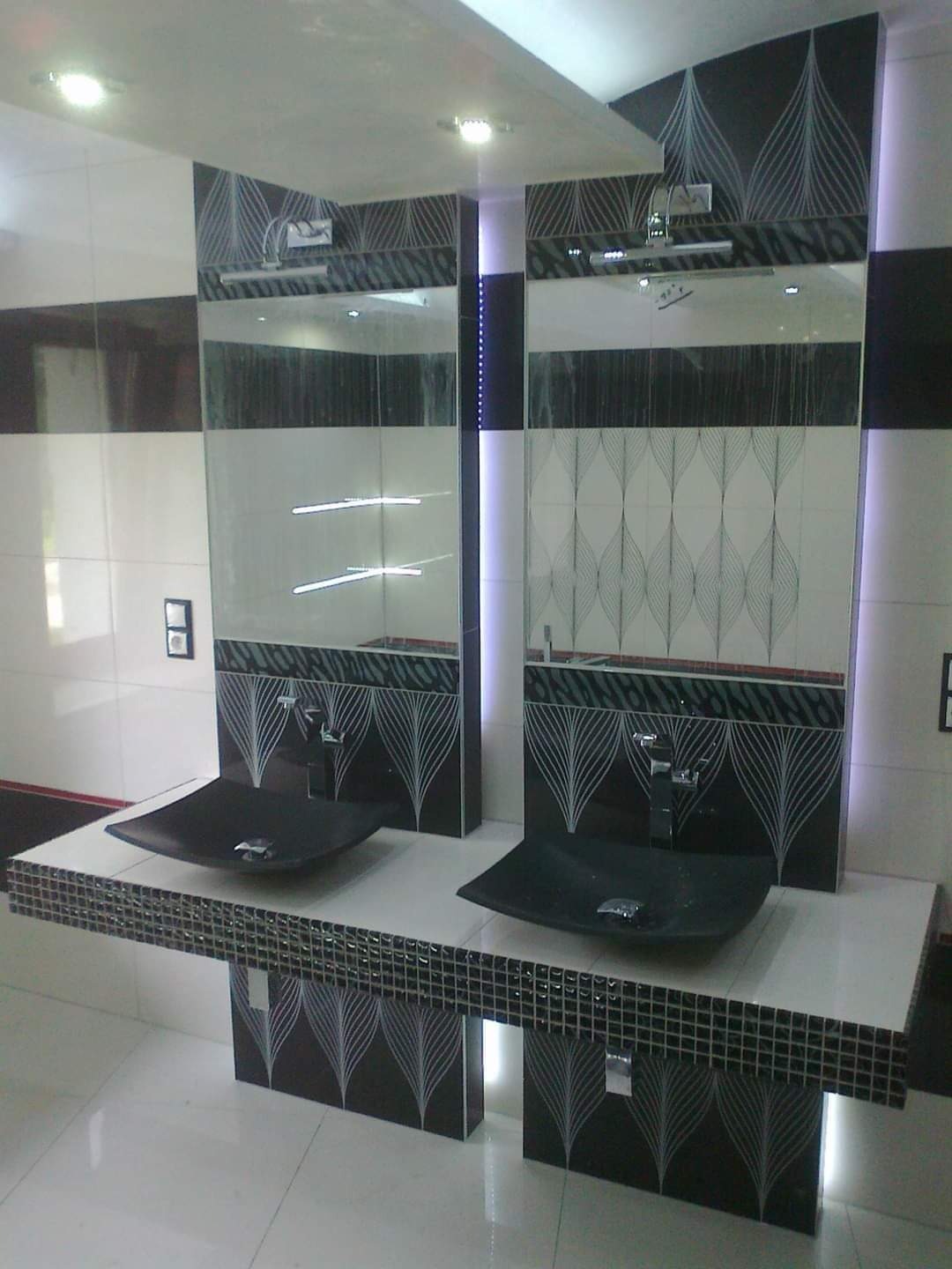 Nowoczesna łazienka z dwoma czarnymi, kwadratowymi umywalkami nablatowymi, dekoracyjną mozaiką i podświetlanymi lustrami z motywem liści.