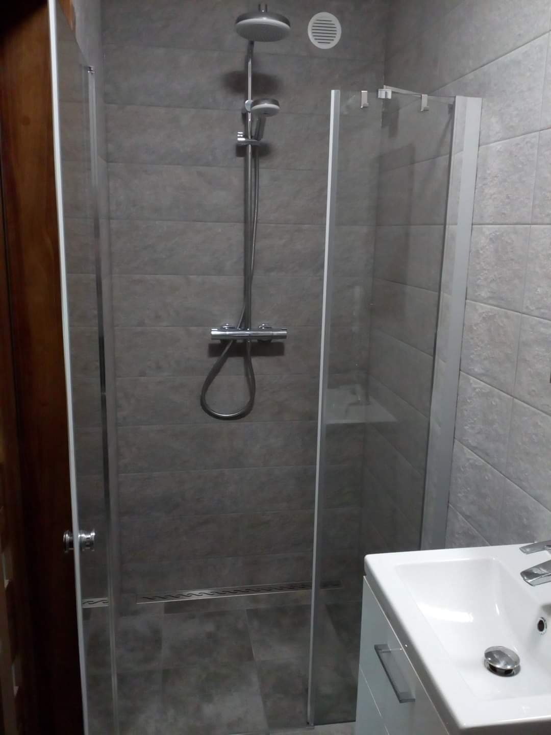 Nowoczesna kabina prysznicowa z szarymi płytkami, deszczownicą i odpływem liniowym, widoczna umywalka z szafką.