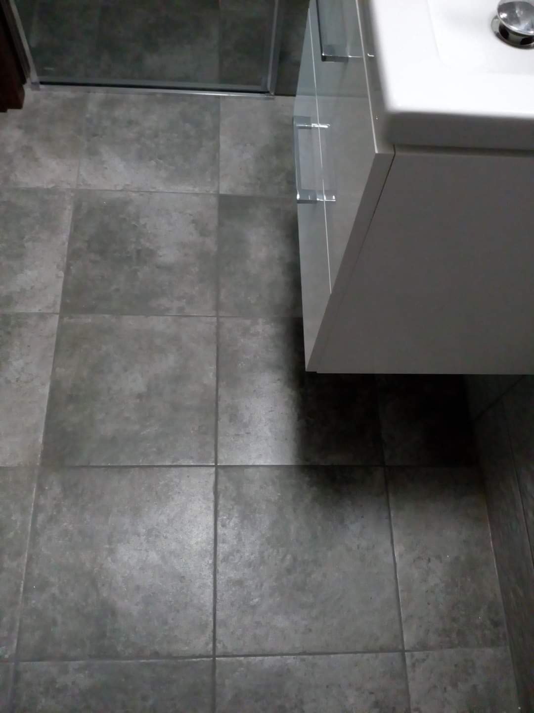 Szare płytki podłogowe w łazience, z widoczną kabiną prysznicową i fragmentem szafki pod umywalką z chromowanymi uchwytami.