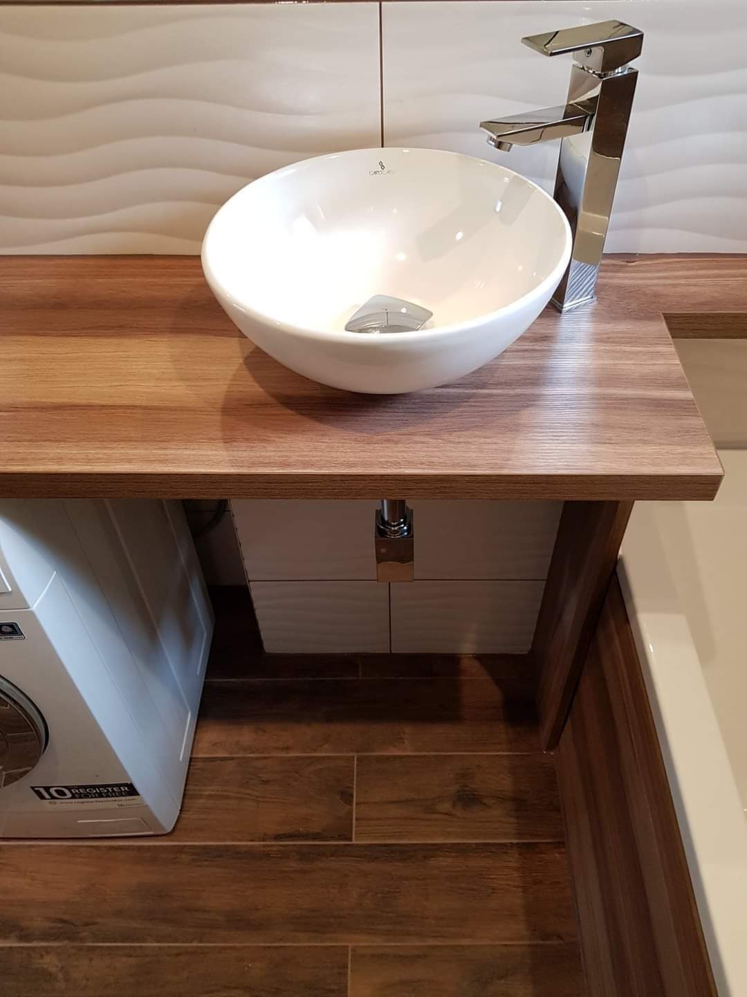 Nowoczesna łazienka z okrągłą umywalką nablatową, chromowaną baterią, drewnianym blatem, płytkami imitującymi drewno na podłodze i ścianie, oraz pralką widoczną z boku.