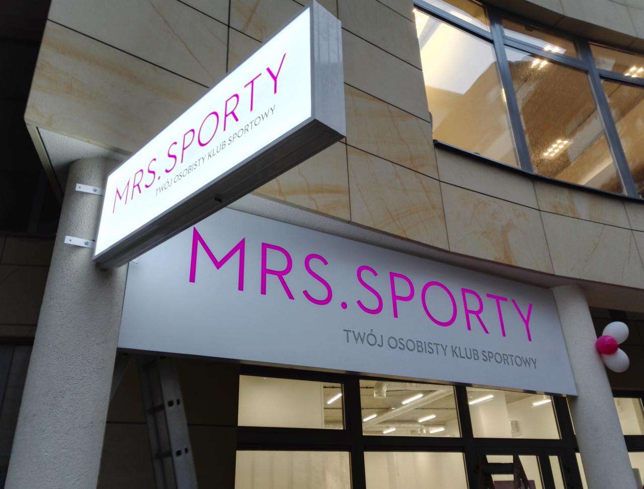 Elewacja budynku z dwoma podświetlanymi szyldami z napisem 'MRS.SPORTY TWÓJ OSOBISTY KLUB SPORTOWY' w kolorze różowym, widoczna drabina i dekoracja balonowa.