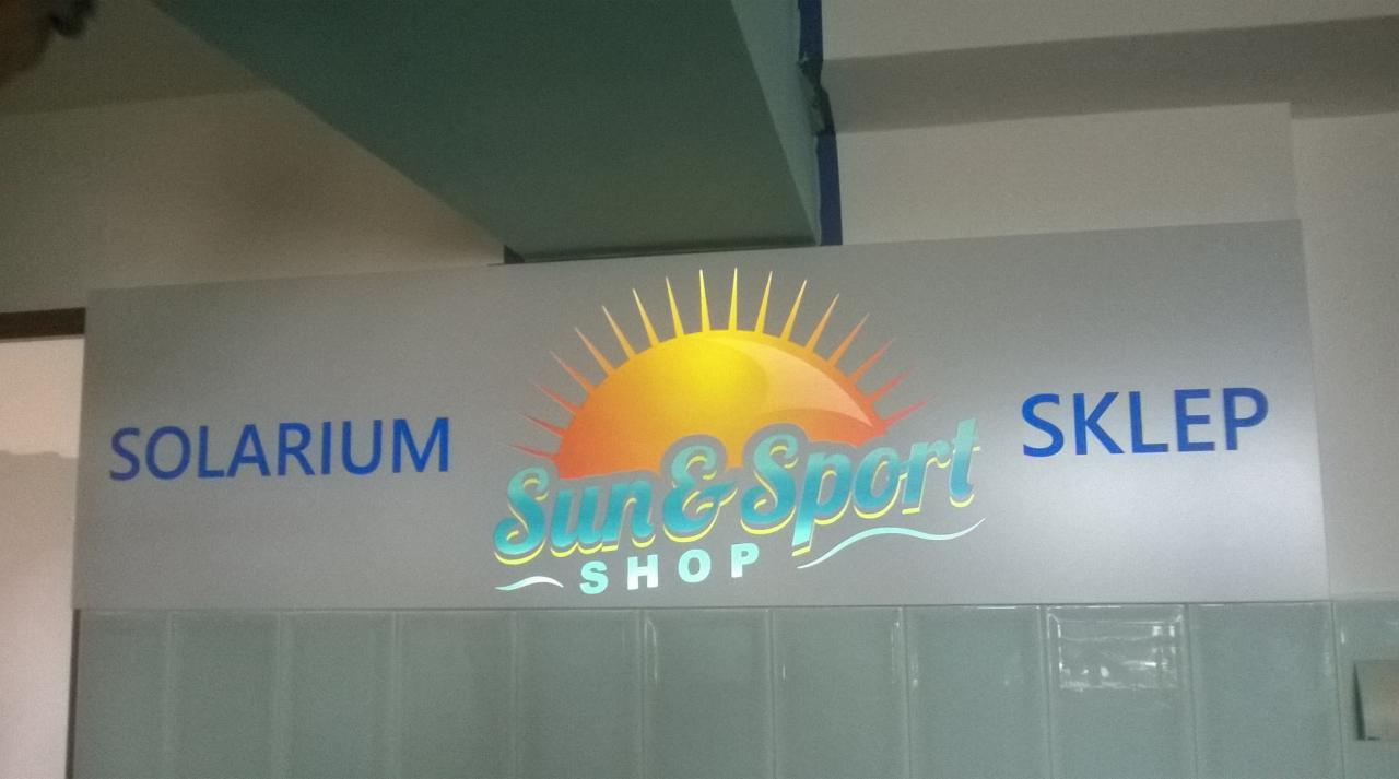 Szyld reklamowy solarium i sklepu Sun & Sport Shop z logo słońca, umieszczony na ścianie z jasnych płytek ceramicznych.