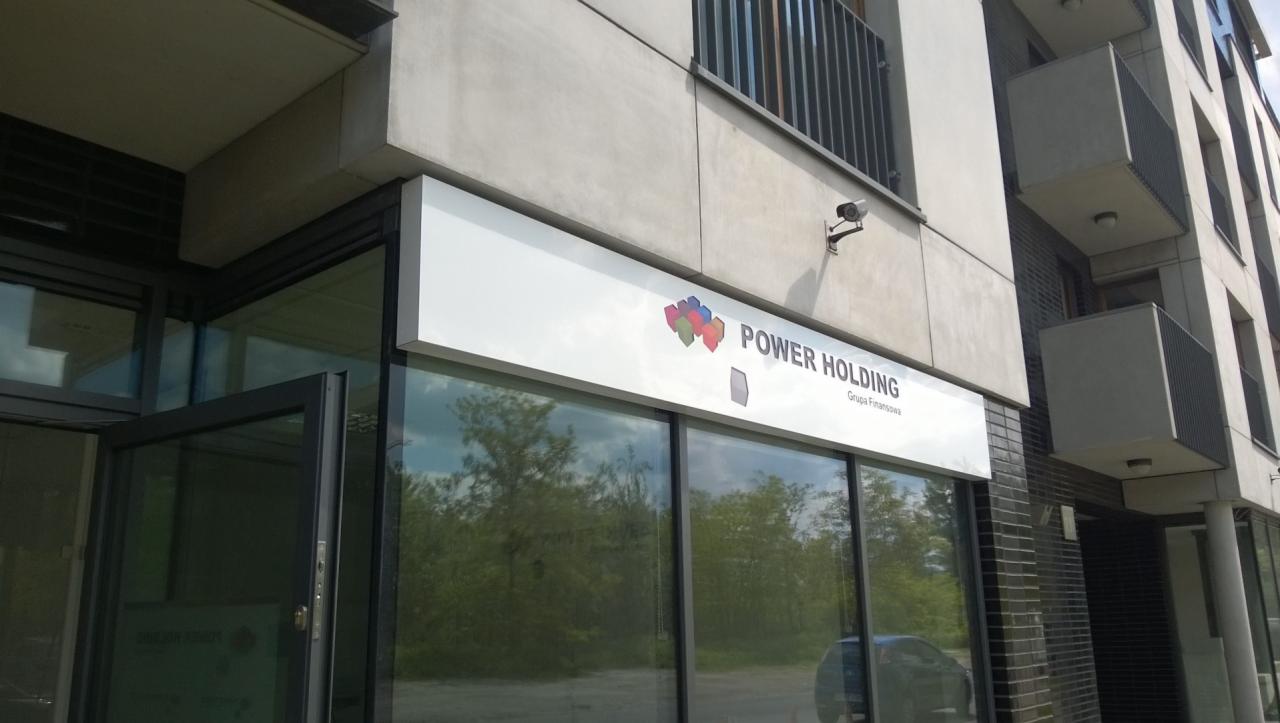 Szyld reklamowy 'Power Holding' nad wejściem do biura, z widocznym logo firmy i odbiciem zieleni w oknach.