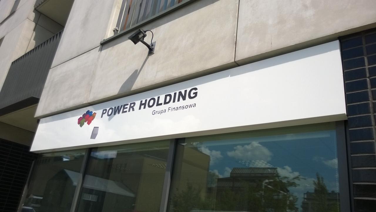 Szyld reklamowy 'Power Holding Grupa Finansowa' nad wejściem do biura, widoczny z zewnątrz, z odbiciem chmur w oknach.