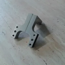 Szlifowany, metalowy element konstrukcyjny z otworami, leżący na marmurowym blacie.