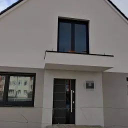 Dwukolorowa elewacja domu jednorodzinnego z czarnymi ramami okiennymi i drzwiami wejściowymi, widok z zewnątrz przez prowizoryczne ogrodzenie.