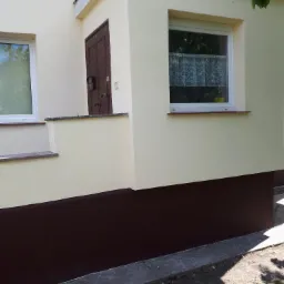 Odnowiona elewacja domu z kremową ścianą i brązowym cokołem, widoczne okna z firankami i drewniane drzwi wejściowe.