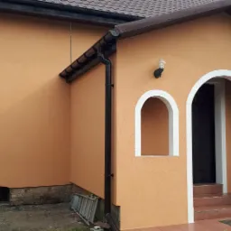 Otynkowana elewacja domu jednorodzinnego w kolorze pomarańczowym z białymi obramowaniami łukowych okien i wejścia, brązowy dach i rynny, widoczne lampy zewnętrzne.