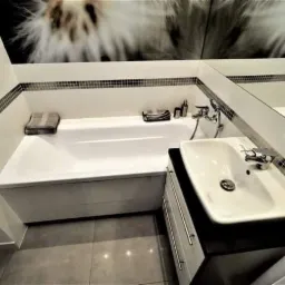 Nowoczesna łazienka z białą wanną obudowaną płytkami, umywalką nablatową na czarnym blacie i mozaiką dekoracyjną wzdłuż górnej krawędzi wanny oraz na ścianach.