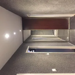 Sufit z szarą, ziarnistą strukturą, w którym zamontowano podłużne okna sufitowe z widokiem na inne pomieszczenia, oraz brązowy panel dekoracyjny z oświetleniem punktowym na ścianie.