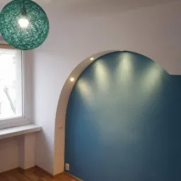 Pomalowany na niebiesko fragment ściany z wnęką oświetloną punktowymi lampami, drewniana podłoga, okno z widocznym fragmentem budynku na zewnątrz oraz zielona lampa wisząca z abażurem z cienkich...