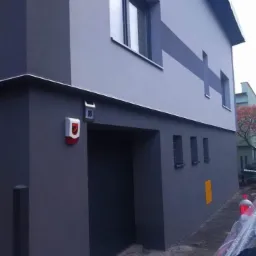 Elewacja dwukondygnacyjnego domu w odcieniach szarości z ciemnoszarą bramą garażową i czerwonym alarmem przy wejściu.