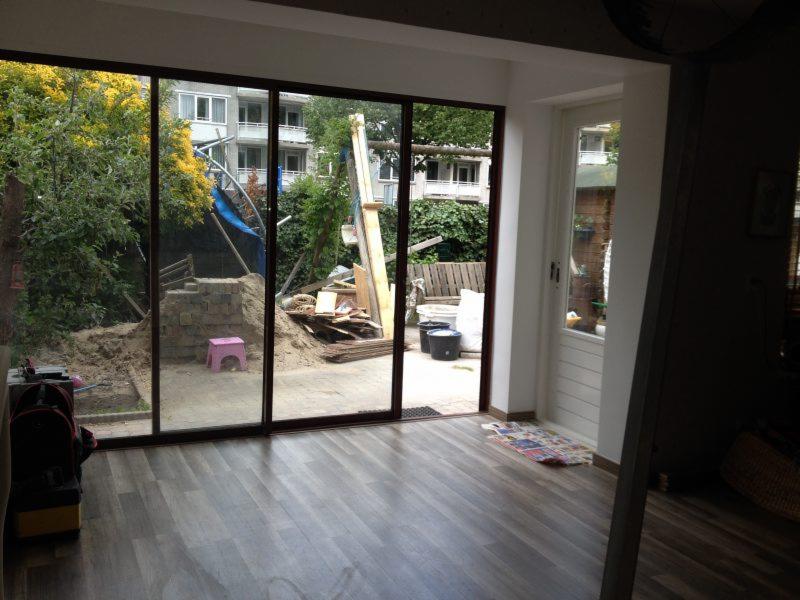 Wnętrze z dużym oknem przesuwnym z widokiem na plac budowy, widoczny różowy stołek, narzędzia i materiały budowlane, podłoga wyłożona panelami.