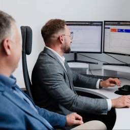 NorEkspert Sp. z o. o. - Mężczyzna w szarej marynarce analizuje dane na kilku monitorach, obok siedzi drugi mężczyzna w niebieskiej marynarce, obaj w biurze z białymi ścianami i biurkiem.