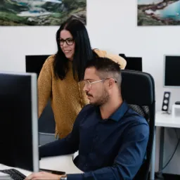 Mężczyzna w okularach pracuje przy komputerze, kobieta w żółtym swetrze stoi za nim i patrzy na ekran, w tle obrazy górskie i jezioro w biurze.