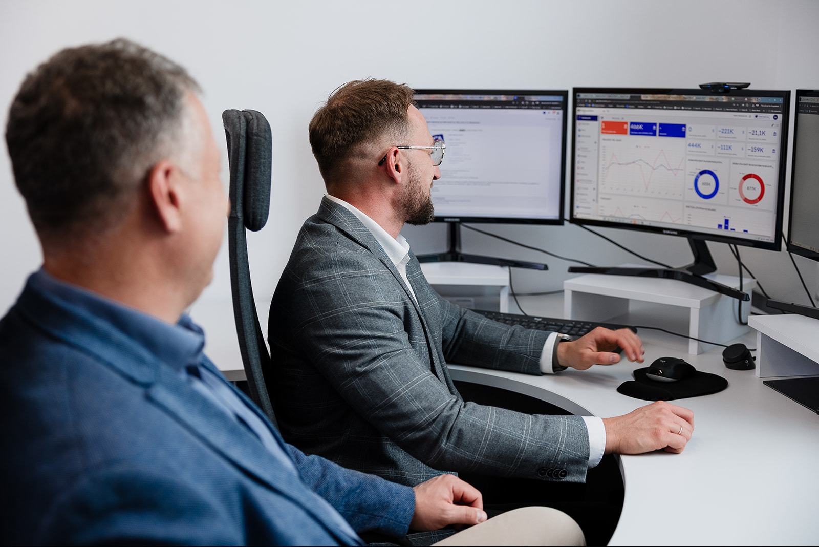 Mężczyzna w szarej marynarce analizuje dane na kilku monitorach, obok siedzi drugi mężczyzna w niebieskiej marynarce, obaj w biurze z białymi ścianami i biurkiem.