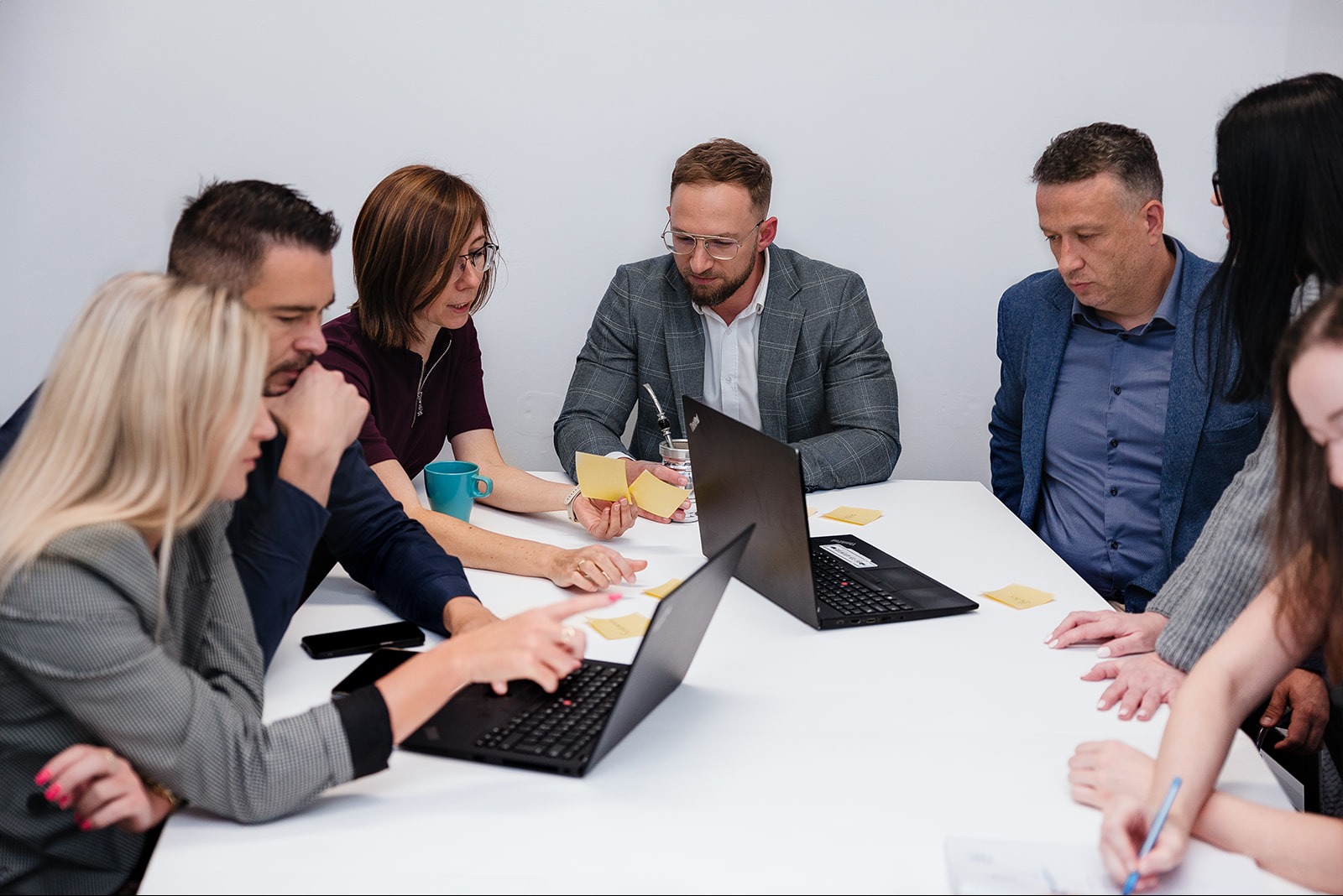 Zespół sześciu osób analizuje notatki i dane na laptopach podczas spotkania przy białym stole.