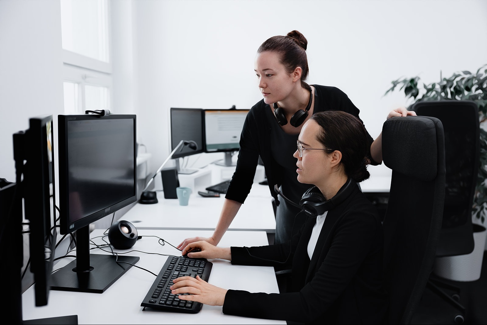 Dwie kobiety w biurze pracują przy komputerach, jedna pisze na klawiaturze, druga stoi za nią i obserwuje ekran, obie mają słuchawki na szyi.