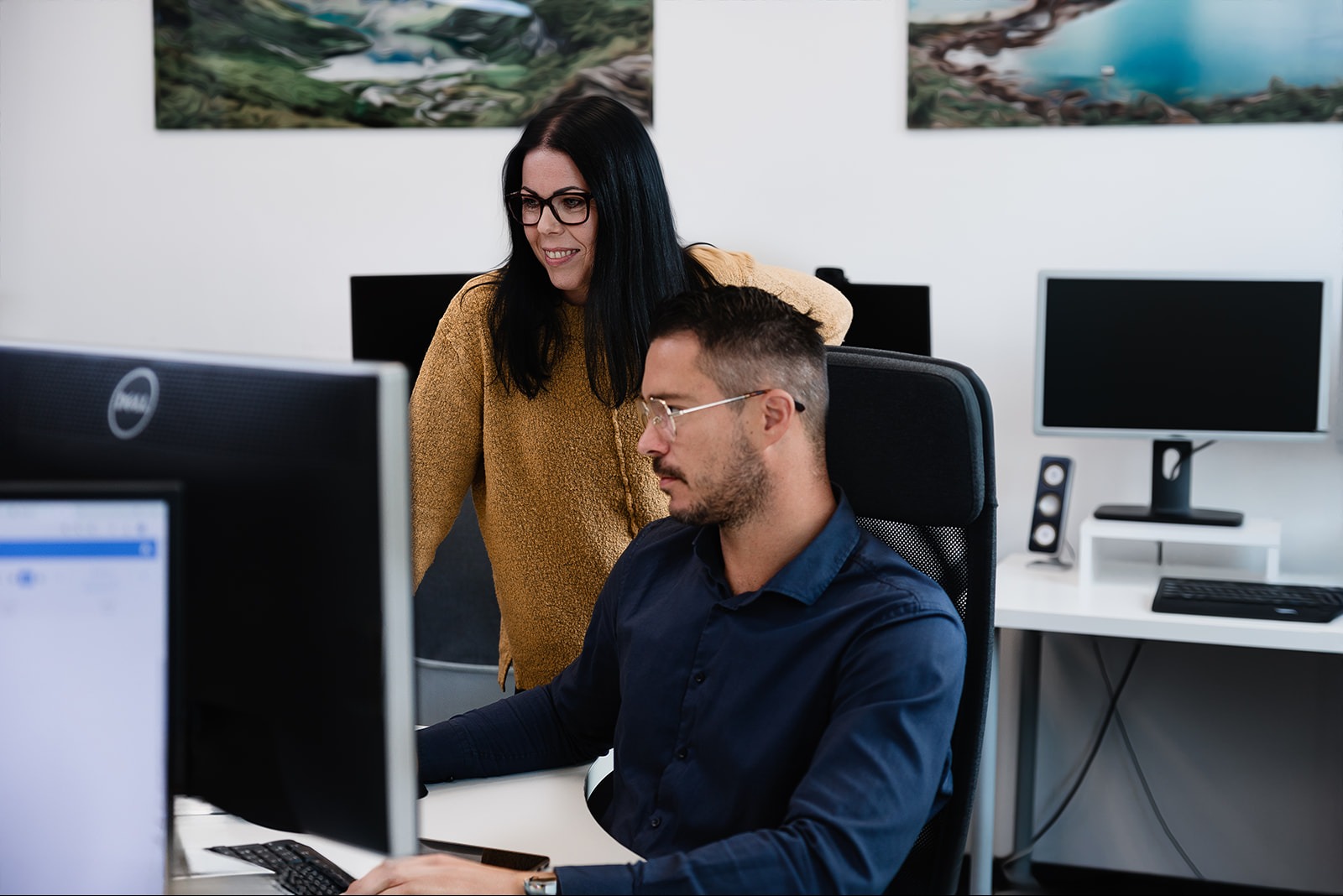 Mężczyzna w okularach pracuje przy komputerze, kobieta w żółtym swetrze stoi za nim i patrzy na ekran, w tle obrazy górskie i jezioro w biurze.