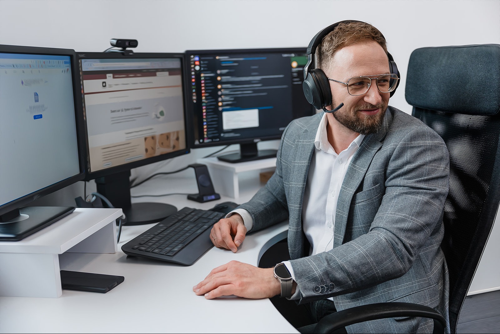 Uśmiechnięty mężczyzna w garniturze i słuchawkach z mikrofonem siedzi przed trzema monitorami, na których widać kody i interfejsy stron internetowych.