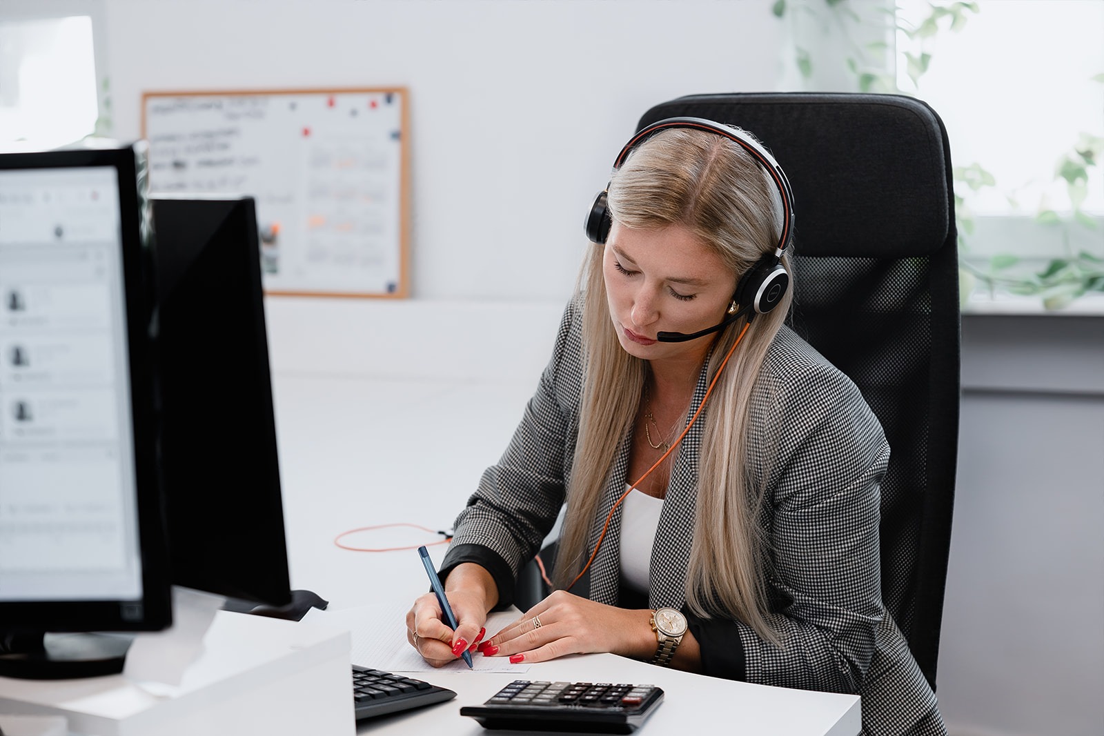 Kobieta w słuchawkach z mikrofonem pisze na kartce przy biurku z komputerem i kalkulatorem, ubrana w szarą marynarkę w kratę i złoty zegarek.