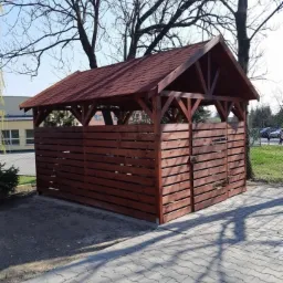 Drewniana konstrukcja z czerwonym dachem, prawdopodobnie wiata śmietnikowa, z poziomymi deskami, zamknięta drzwiczkami z zasuwą, na kostce brukowej, w tle drzewa i budynek.