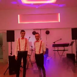 Dwaj mężczyźni w białych koszulach i czerwonych muchach oraz szelkach stoją przed sprzętem muzycznym w pomieszczeniu oświetlonym na różowo z efektem dymu.