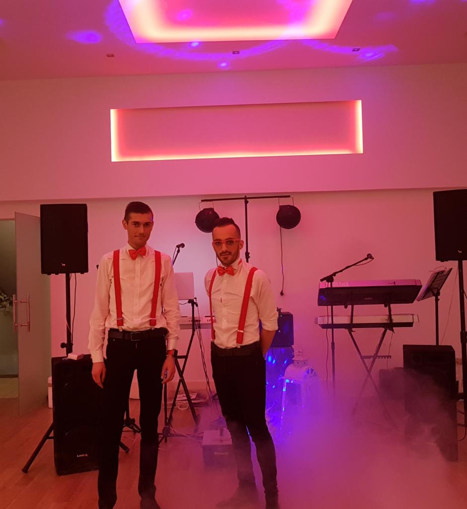 Dwaj mężczyźni w białych koszulach i czerwonych muchach oraz szelkach stoją przed sprzętem muzycznym w pomieszczeniu oświetlonym na różowo z efektem dymu.