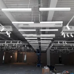 Wnętrze sklepu w trakcie prac wykończeniowych, widoczne sufitowe instalacje oświetleniowe: szyny z reflektorami punktowymi i podłużne lampy LED, podnośnik koszowy na podłodze.