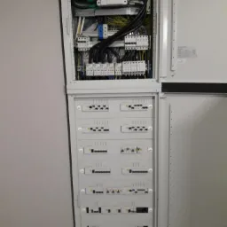 Otwarta szafa elektryczna z widocznymi bezpiecznikami, przewodami i elementami sterującymi w szarej obudowie, zamontowana na jasnej ścianie.