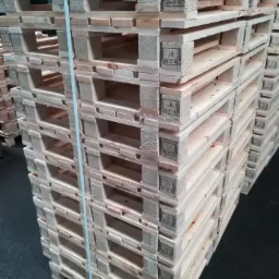 Stos drewnianych palet euro, zabezpieczonych niebieską taśmą, z widocznymi oznaczeniami fitosanitarnymi HT, ustawionych na ciemnej posadzce w magazynie.