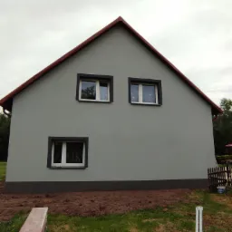 Świeżo pomalowana szara elewacja domu jednorodzinnego z brązowymi ramami okiennymi i dachem, widoczne ślady prac ziemnych wok&oacute;ł budynku.