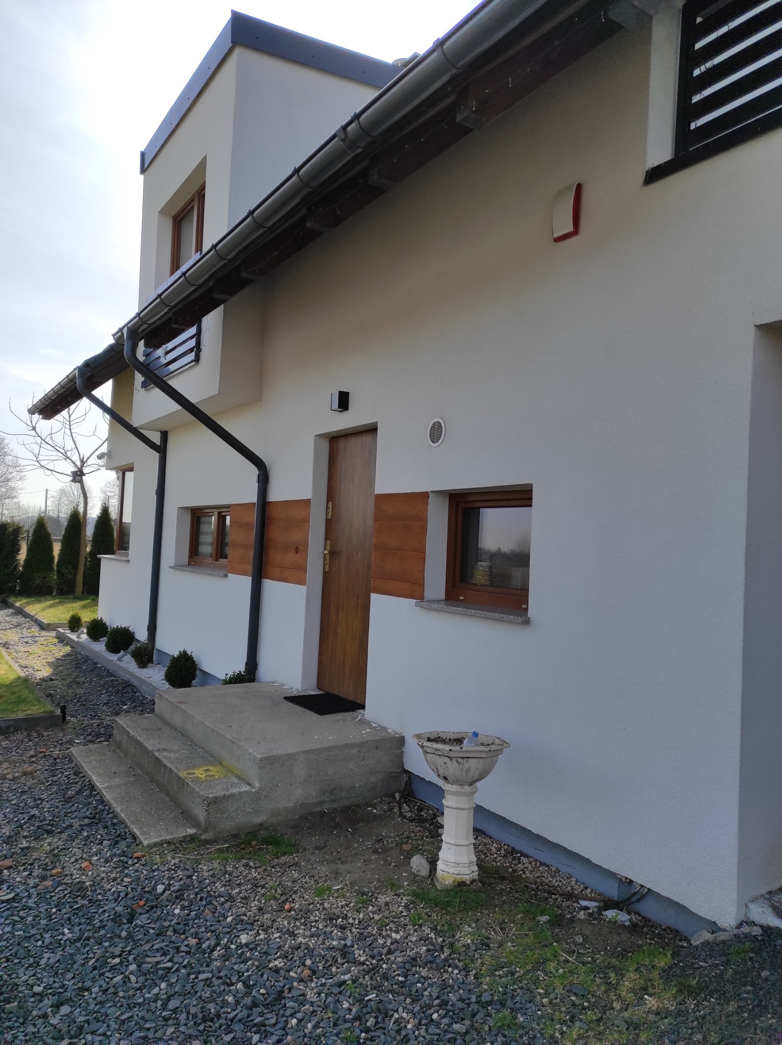 Nowoczesny dom jednorodzinny z białą elewacją, drewnianymi akcentami na drzwiach i oknach, oraz czarnymi rynnami. Widoczne betonowe schody wejściowe i kamienny podjazd.