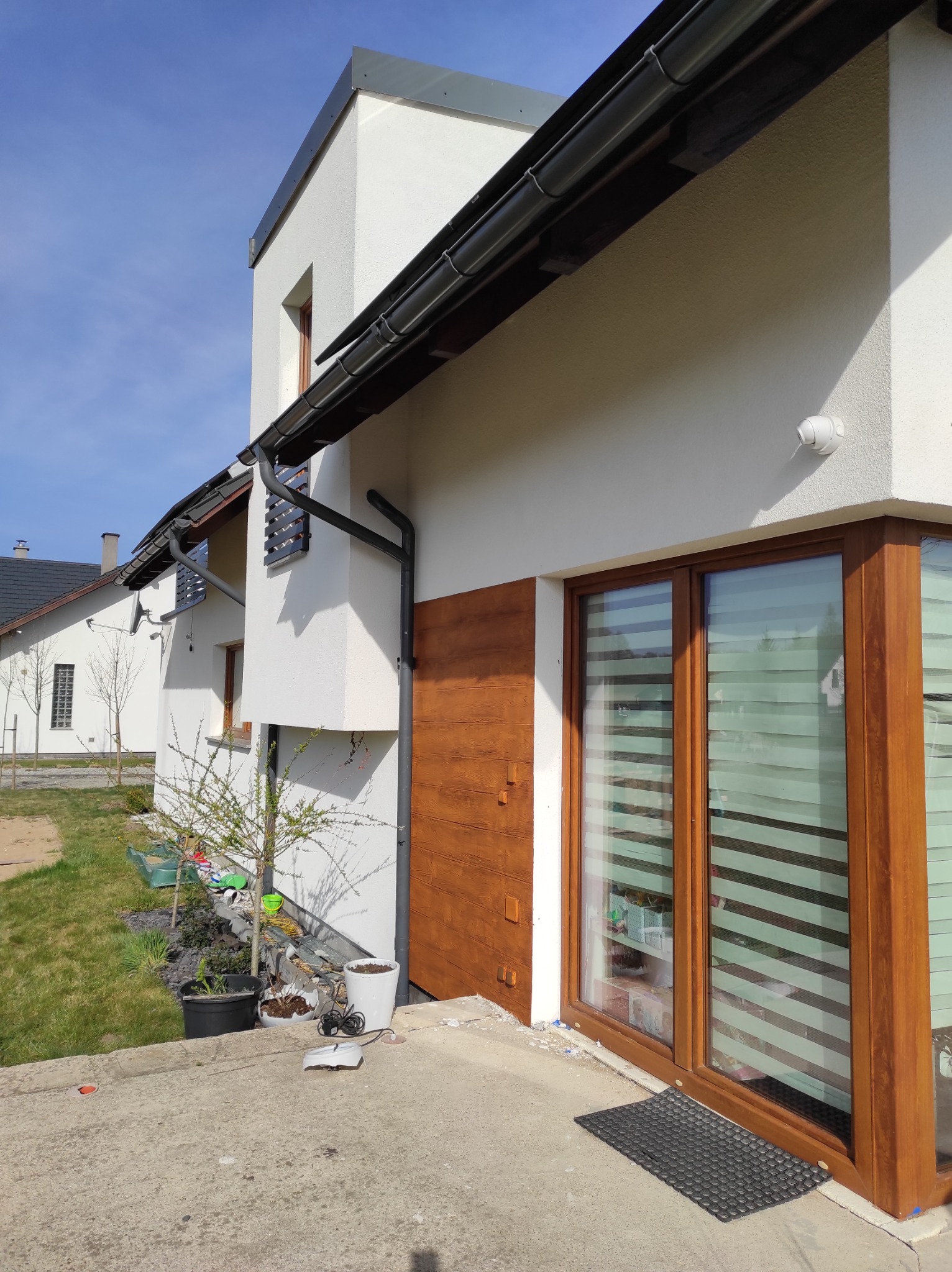 Nowoczesny dom z białą elewacją, drewnianą okładziną i dużymi oknami z roletami, widok z zewnątrz na trawnik i rośliny w donicach.