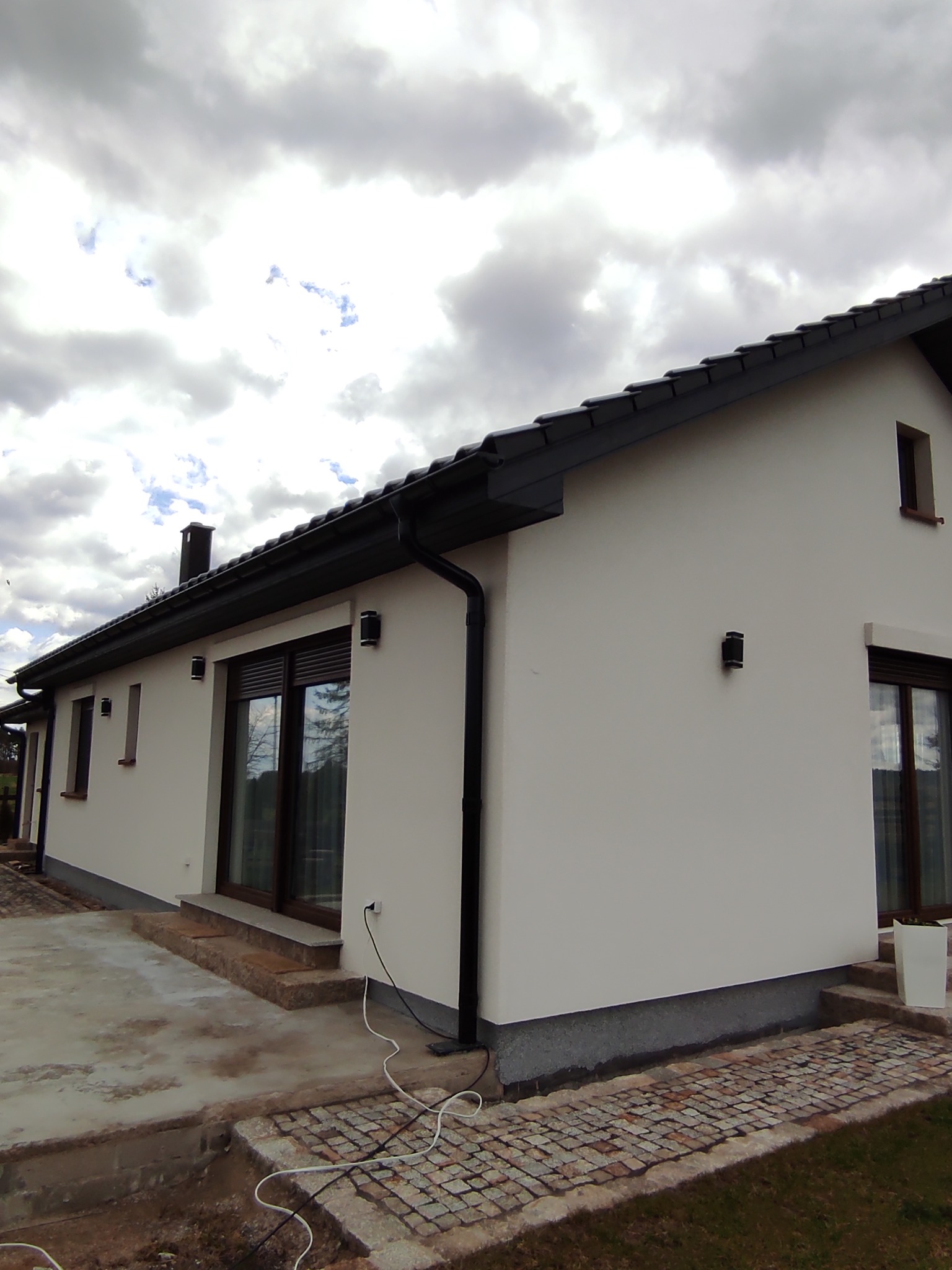 Jednopiętrowy dom z jasną, gładką elewacją i ciemną dachówką, widoczny fragment brukowanego chodnika i niedokończone prace ziemne wokół budynku.