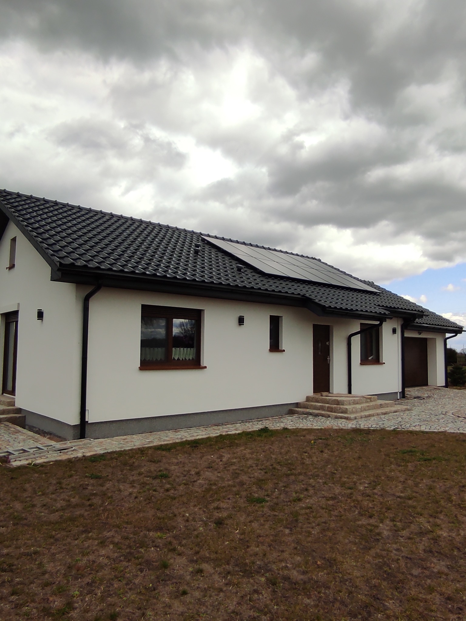 Biały dom z panelami słonecznymi na czarnym dachu z dachówki, widok z boku, otoczony brukowaną ścieżką i trawnikiem przed pochmurnym niebem.