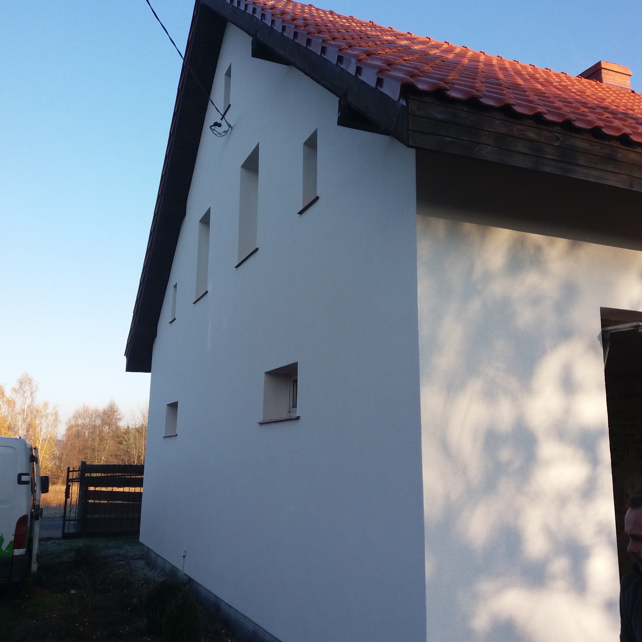 Wykończona elewacja budynku jednorodzinnego z nową, czerwoną dachówką, widoczne prostokątne okna i fragment zaparkowanego samochodu dostawczego.