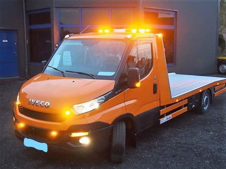 Pomarańczowa laweta Iveco z włączonymi światłami ostrzegawczymi, zaparkowana przed budynkiem z ciemnymi oknami.