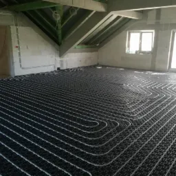 Rozłożona instalacja ogrzewania podłogowego w surowym stanie, rury ułożone na czarnej macie, widoczne okna i surowy beton.