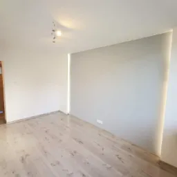 Pomalowane na szaro puste wnętrze pokoju z panelami podłogowymi, oświetleniem szynowym i pionowym paskiem świetlnym w rogu ściany.