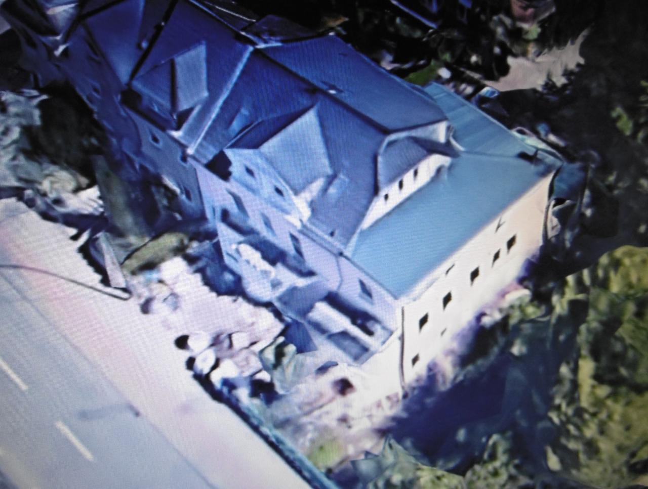 Widok z góry na budynek z ciemnym dachem i jasną elewacją, otoczony zielenią i fragmentem drogi, z widocznymi oknami w rzędzie.