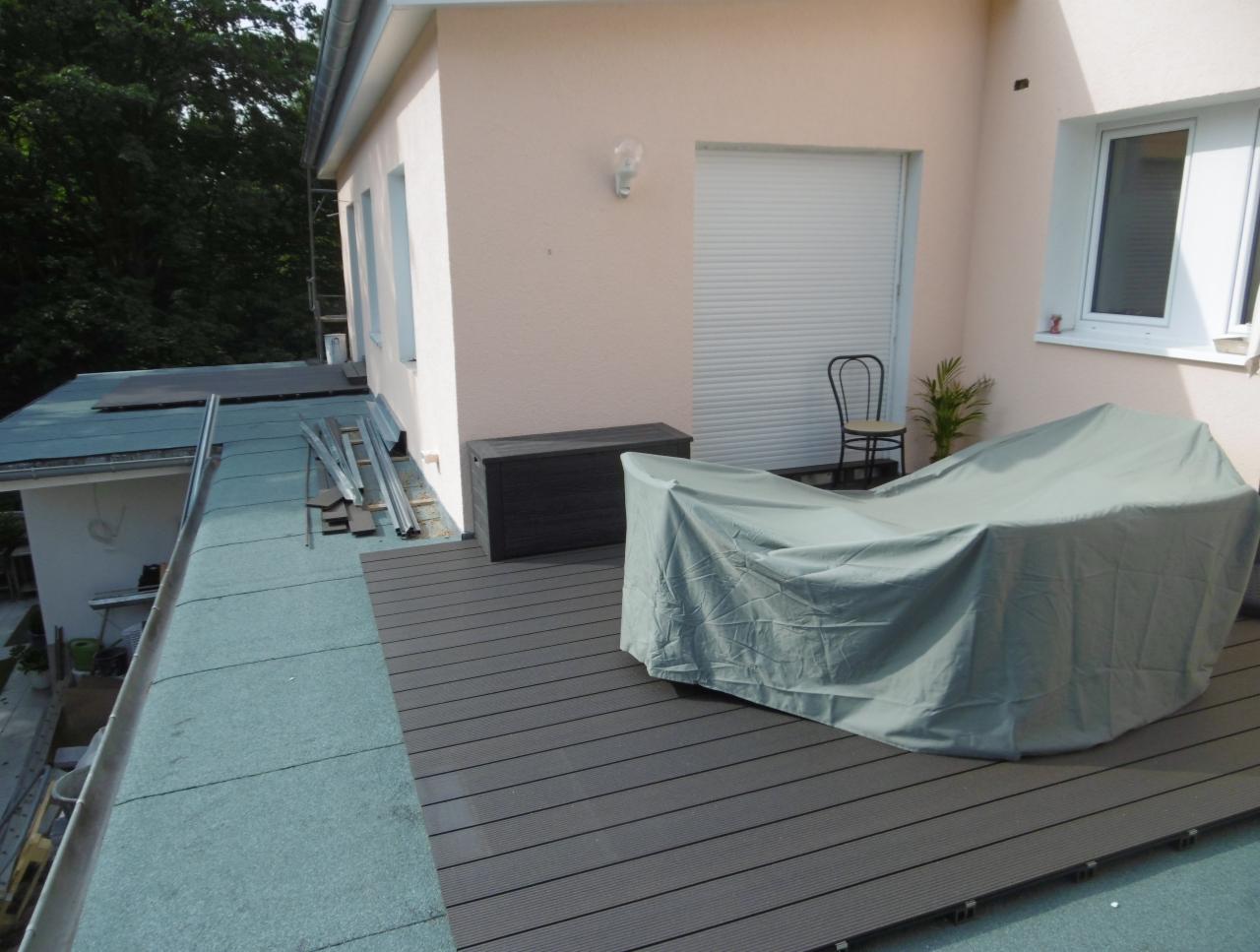 Taras na dachu z kompozytowych desek w kolorze grafitowym, częściowo zadaszony, meble ogrodowe przykryte pokrowcem, widoczne materiały budowlane i narzędzia na przyległym dachu.