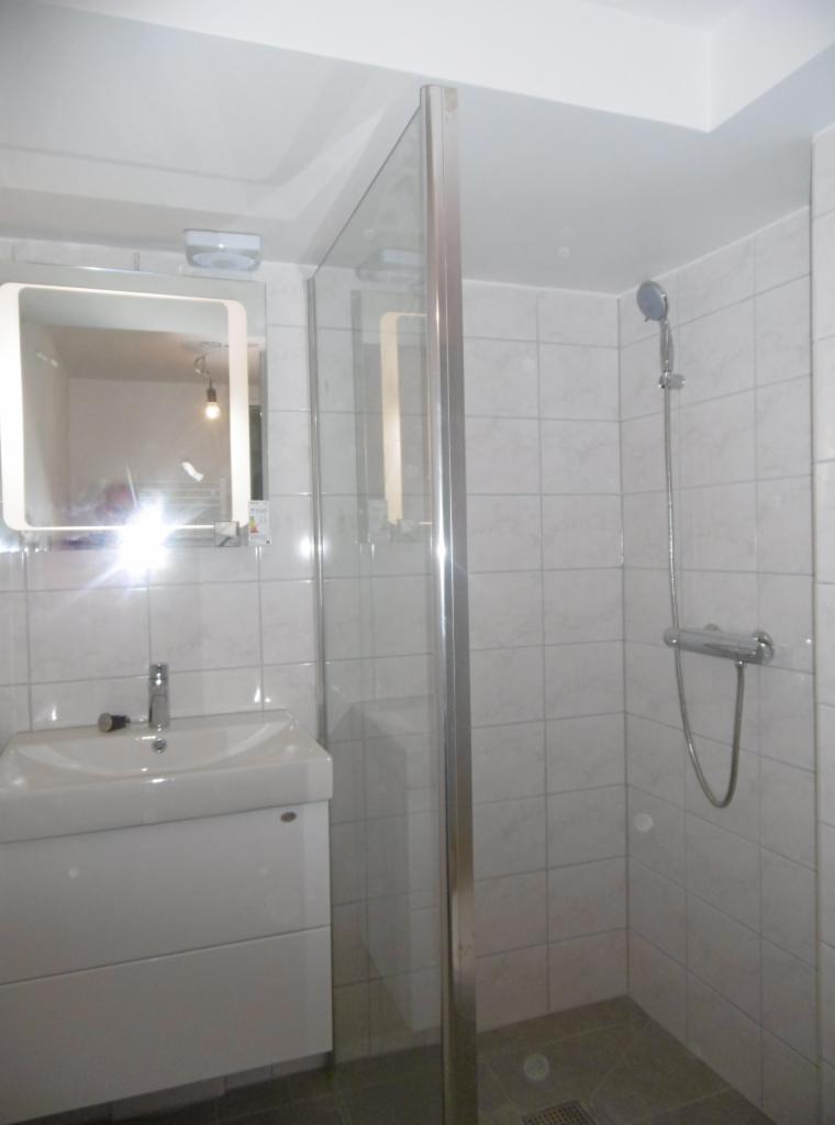 Biała łazienka z kabiną prysznicową ze szklaną ścianką, umywalką z szafką i lustrem z oświetleniem, wykończona kwadratowymi płytkami w odcieniach szarości.