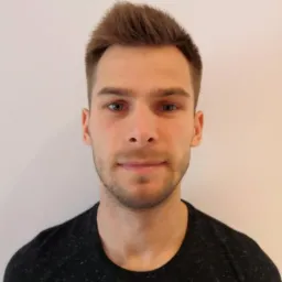 Portret młodego mężczyzny z krótkimi brązowymi włosami i lekkim zarostem na tle jasnej ściany, ubrany w ciemną koszulkę. Ujęcie od przodu, skupione na twarzy.