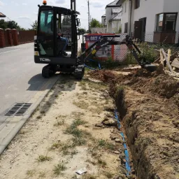 Czarna mini koparka TC16-2 kopie wąski rów w piaszczystej ziemi, w którym ułożono niebieskie rury. W tle widoczny budynek mieszkalny i ogrodzenie z siatki.