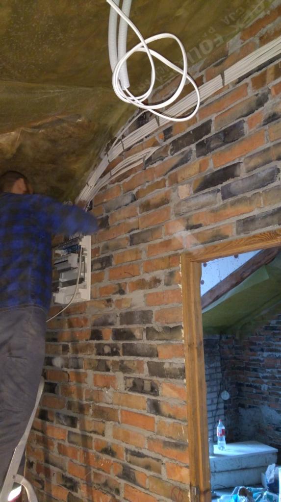 Elektryk na drabinie montuje rozdzielnicę elektryczną na ceglanej ścianie w pomieszczeniu z surowym wykończeniem, widoczne kable w peszlach i izolacja stropu.