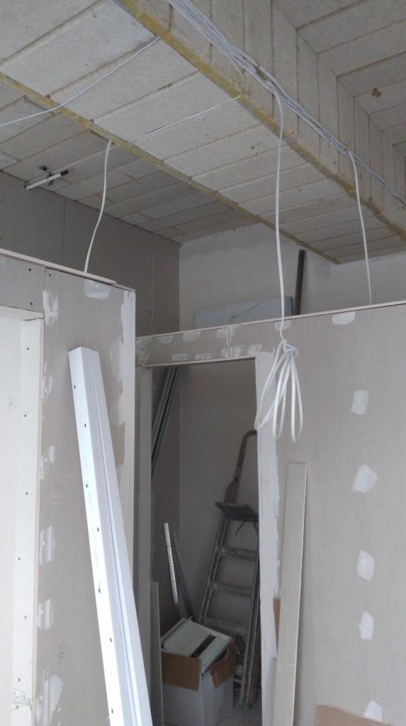 Wnętrze w trakcie remontu: widoczne kable elektryczne pod sufitem, częściowo zamontowane płyty gipsowo-kartonowe na ścianach, drabina i materiały budowlane w tle.