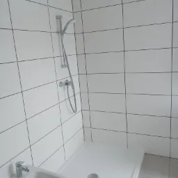 Nowoczesna, minimalistyczna łazienka z białymi, kwadratowymi płytkami, kabiną prysznicową bez brodzika i bidetem z chromowaną armaturą.