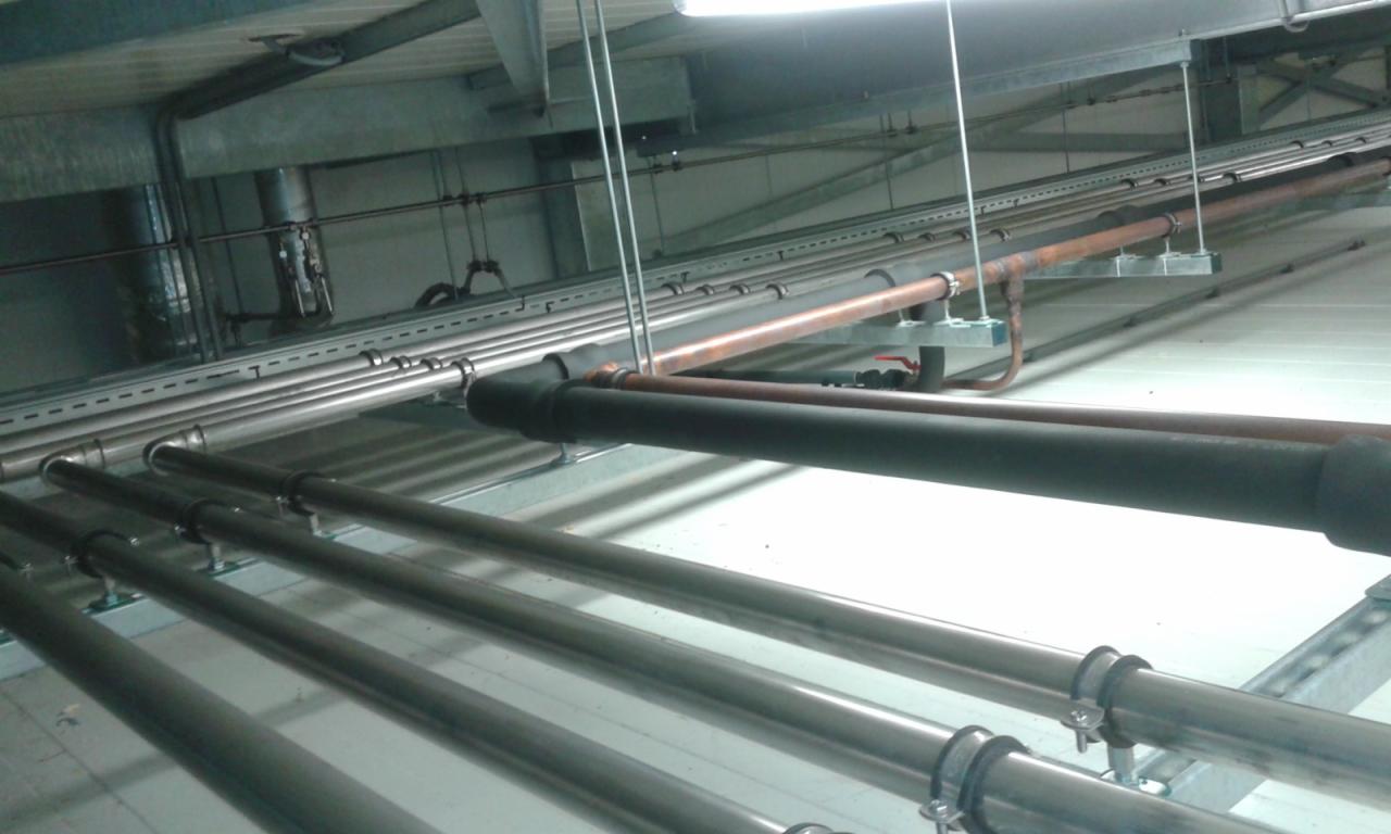 System rur stalowych i miedzianych, zamocowanych pod sufitem w hali, z widoczną izolacją na jednej z rur.